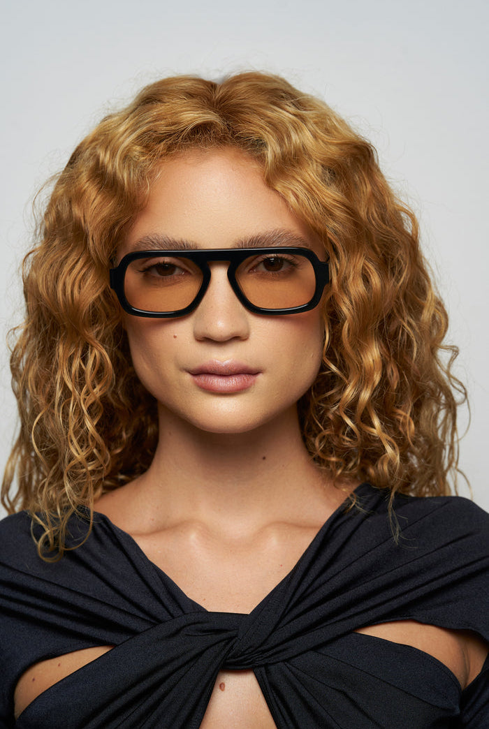 Woman wearing black glasses and a black top with a bow, against a light gray background