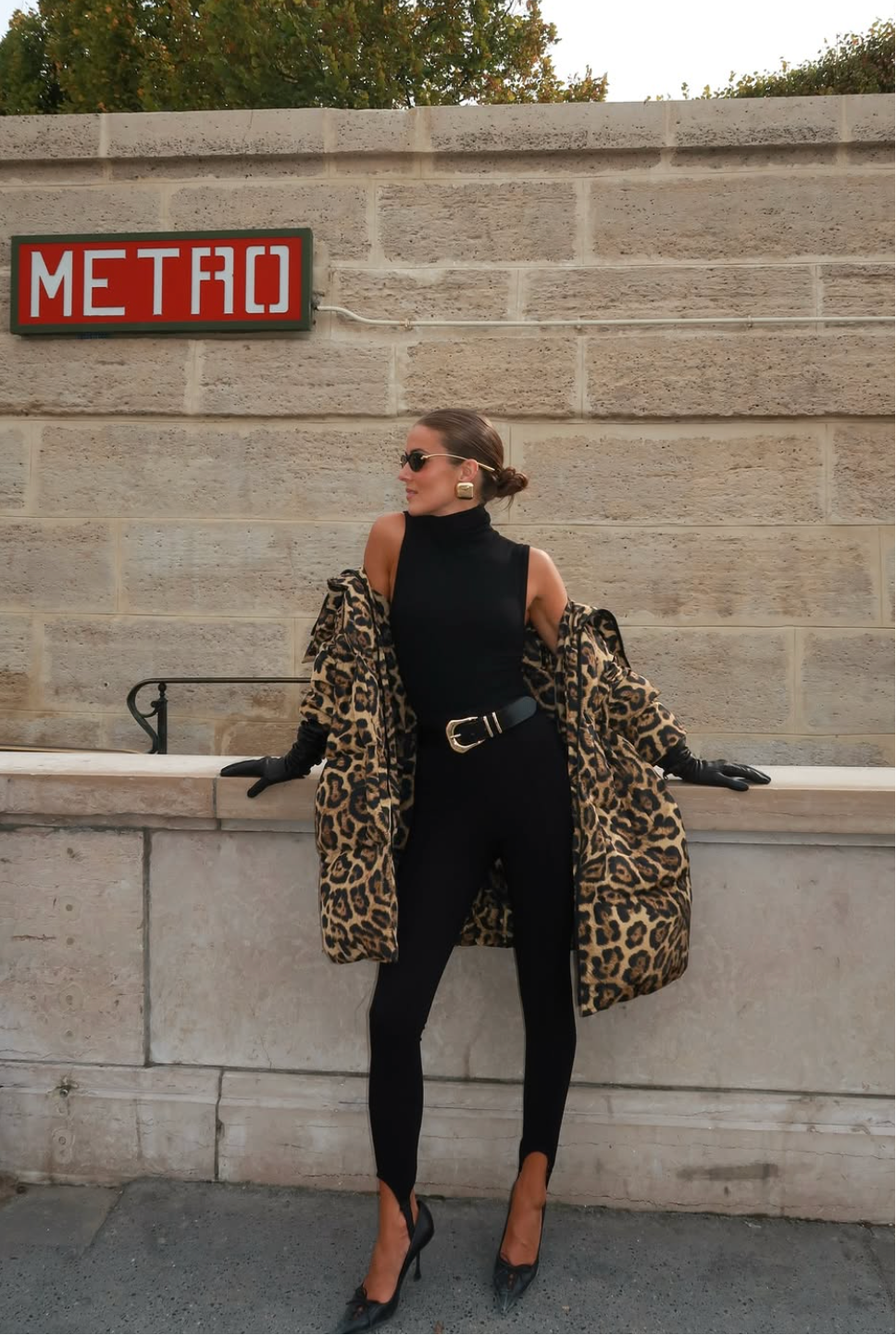 Woman in black outfit with leopard print coat standing against a stone wall with a 'METRO' sign.